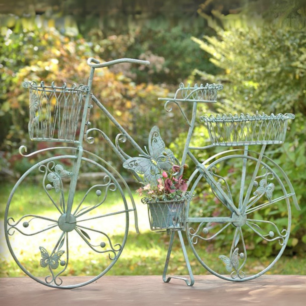 A decorative metal garden sculpture shaped like a vintage bicycle, painted in a distressed light green/teal finish, with butterfly details and a small flower basket on the front, set outdoors on a wooden surface with a blurred garden background.