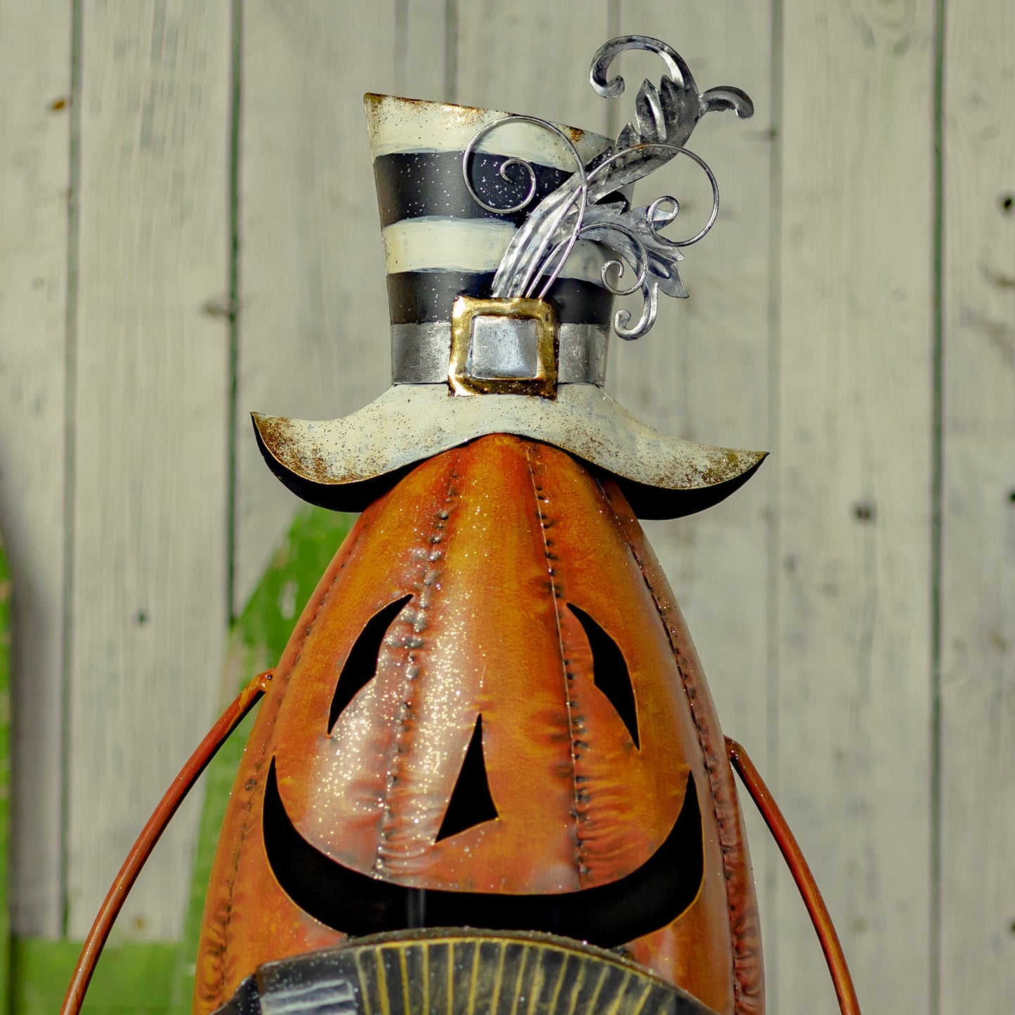 A decorative metal pumpkin sculpture with a carved jack-o'-lantern face, topped with a black-and-white striped hat and silver scroll accents, photographed against a light wood plank background.