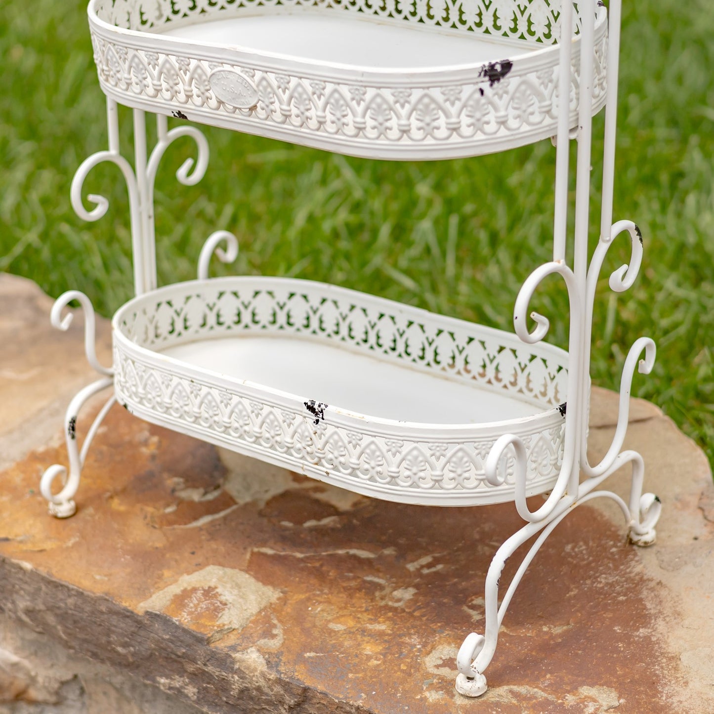 A white metal two-tier garden shelf with ornate scrollwork and cutout detailing, shown outdoors on a stone surface with green grass in the background.