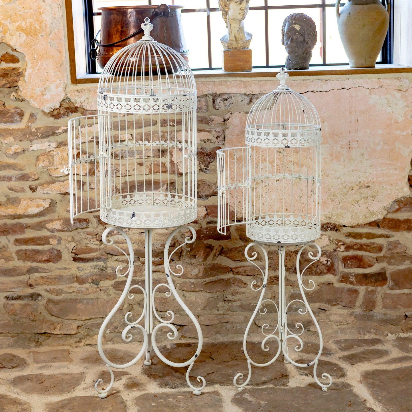 Two white ornate metal birdcage stands with open doors, displayed against a rustic stone wall and window backdrop.