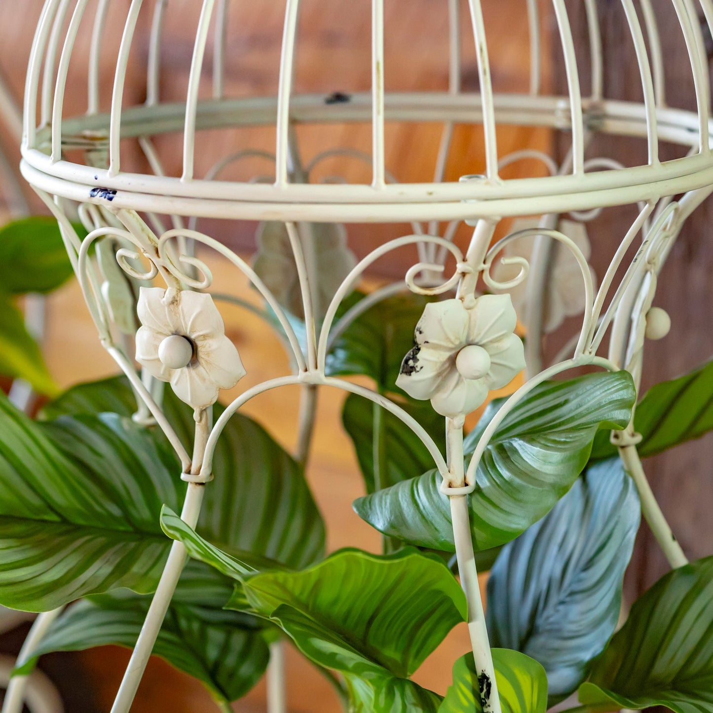 An off-white metal birdcage-style plant stand with decorative scrollwork and flower details, holding green foliage against a softly blurred warm brown background.