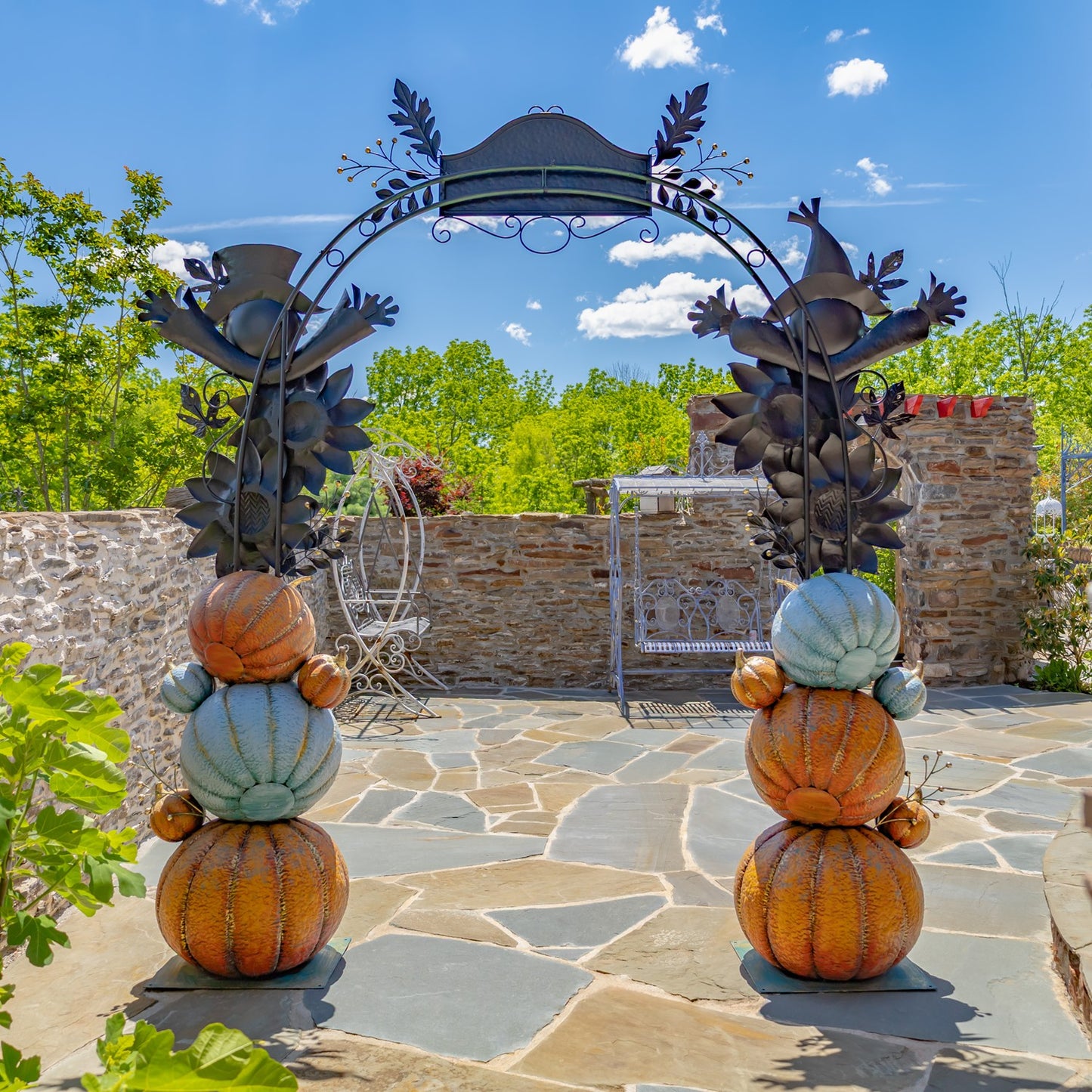 Outdoor autumn garden arch decoration with stacked faux pumpkins and gourds in orange, tan, and pale blue, flanked by dark metal leafy gate supports on a stone patio under a blue sky.