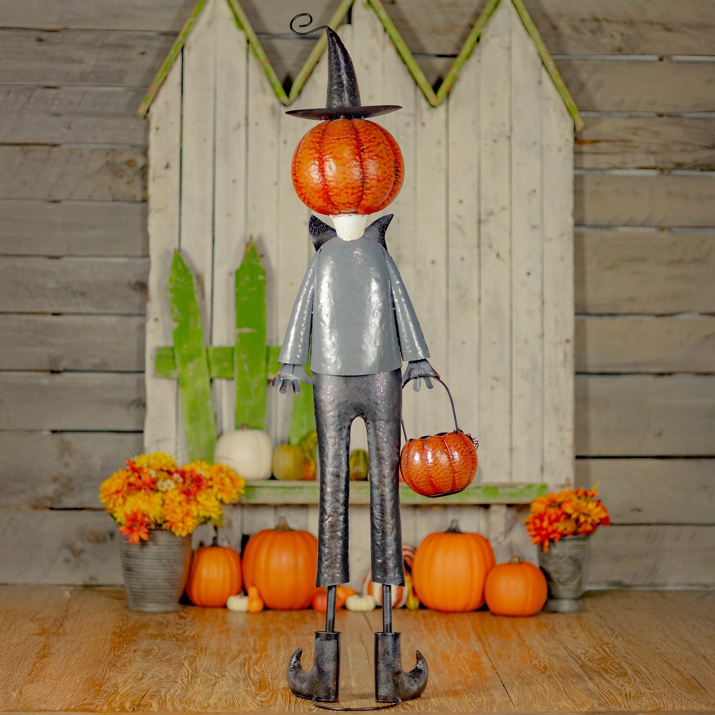 Decorative figure with a pumpkin head and bucket, standing on a wooden floor with pumpkins and flowers.