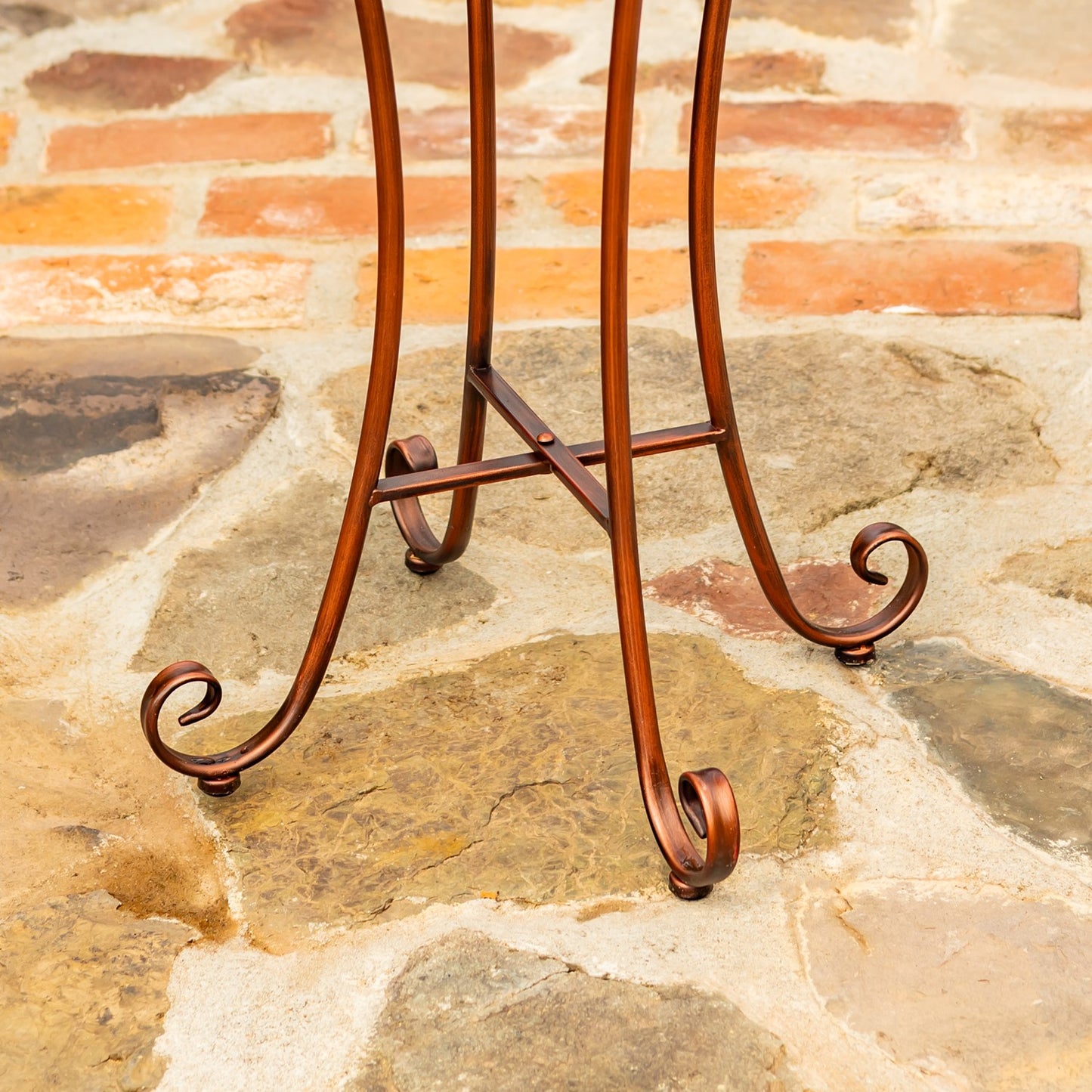 A wrought iron plant stand with a warm copper-brown finish, featuring elegant curved legs and scrollwork feet, set on a rustic stone patio with a brick wall background.