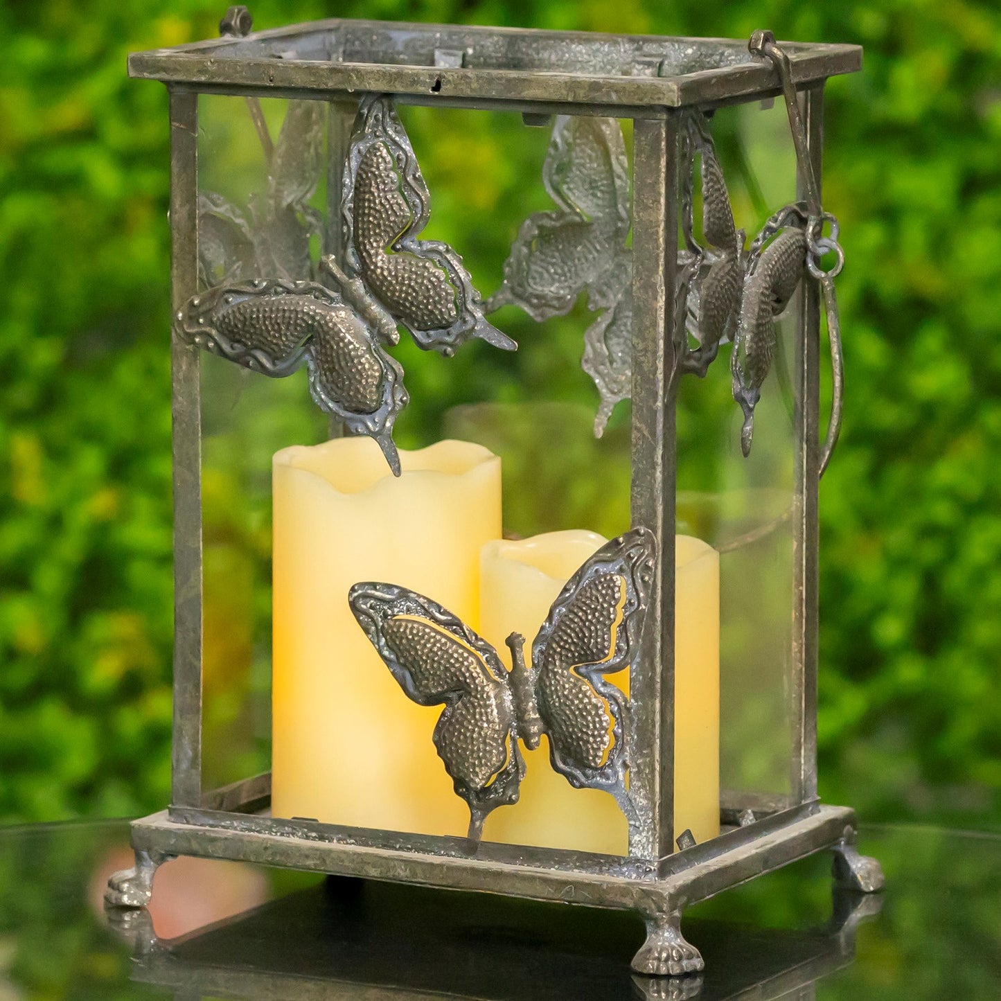 A decorative metal lantern with butterfly cutouts and two lit cream candles inside, photographed outdoors against a blurred green background.