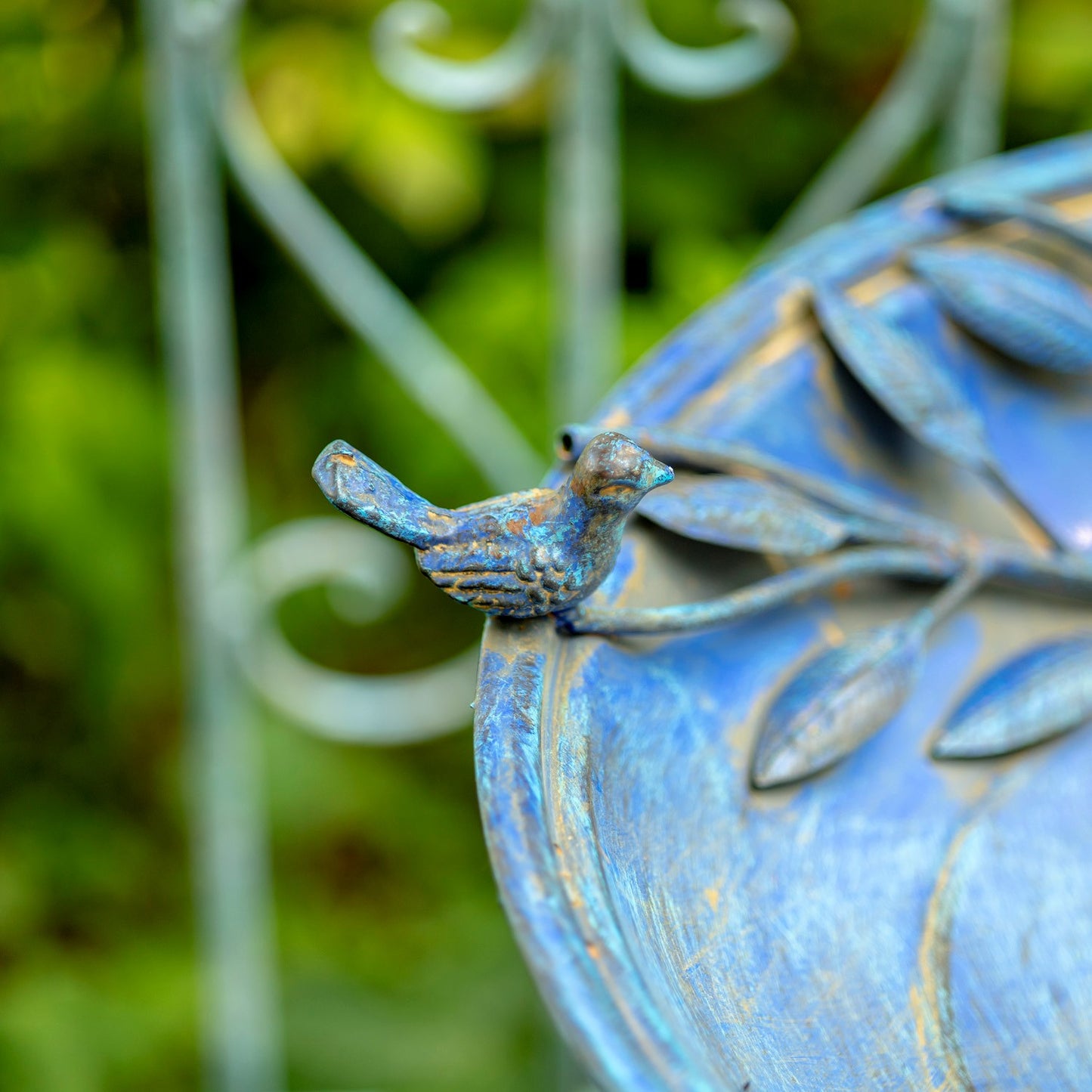 A close-up shot of a small bird figurine perched on a blue metal garden bench with leaf designs, set against a blurred green garden background.