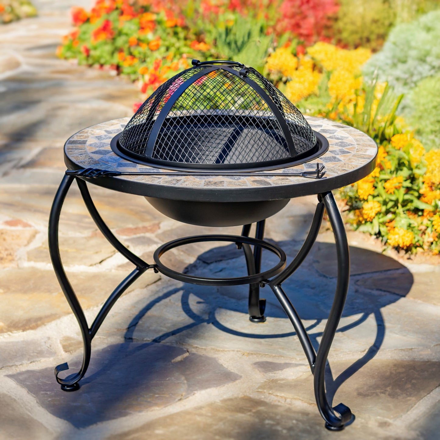 A round black metal fire pit table with a tiled top and a dome-shaped fire guard, shown outdoors on a stone patio with colorful flowers in the background.