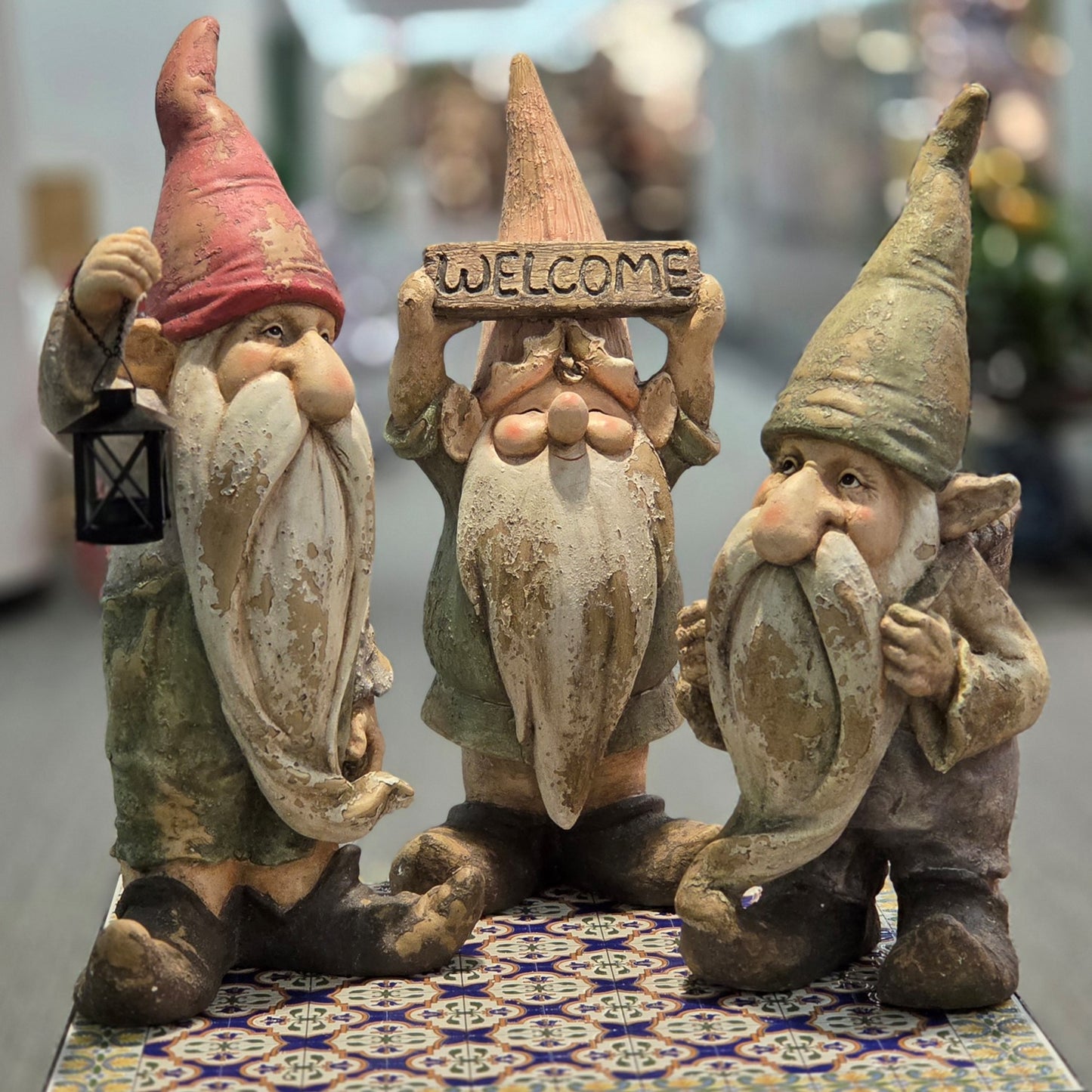 Three rustic garden gnome figurines with long beards, displayed together on a patterned tile surface. One gnome holds a lantern, the center gnome holds a wooden sign reading 'WELCOME', and the right gnome holds a small object near its face.