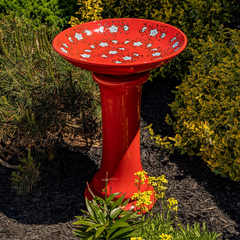 Red porcelain birdbath with blue flowers in a garden scene.