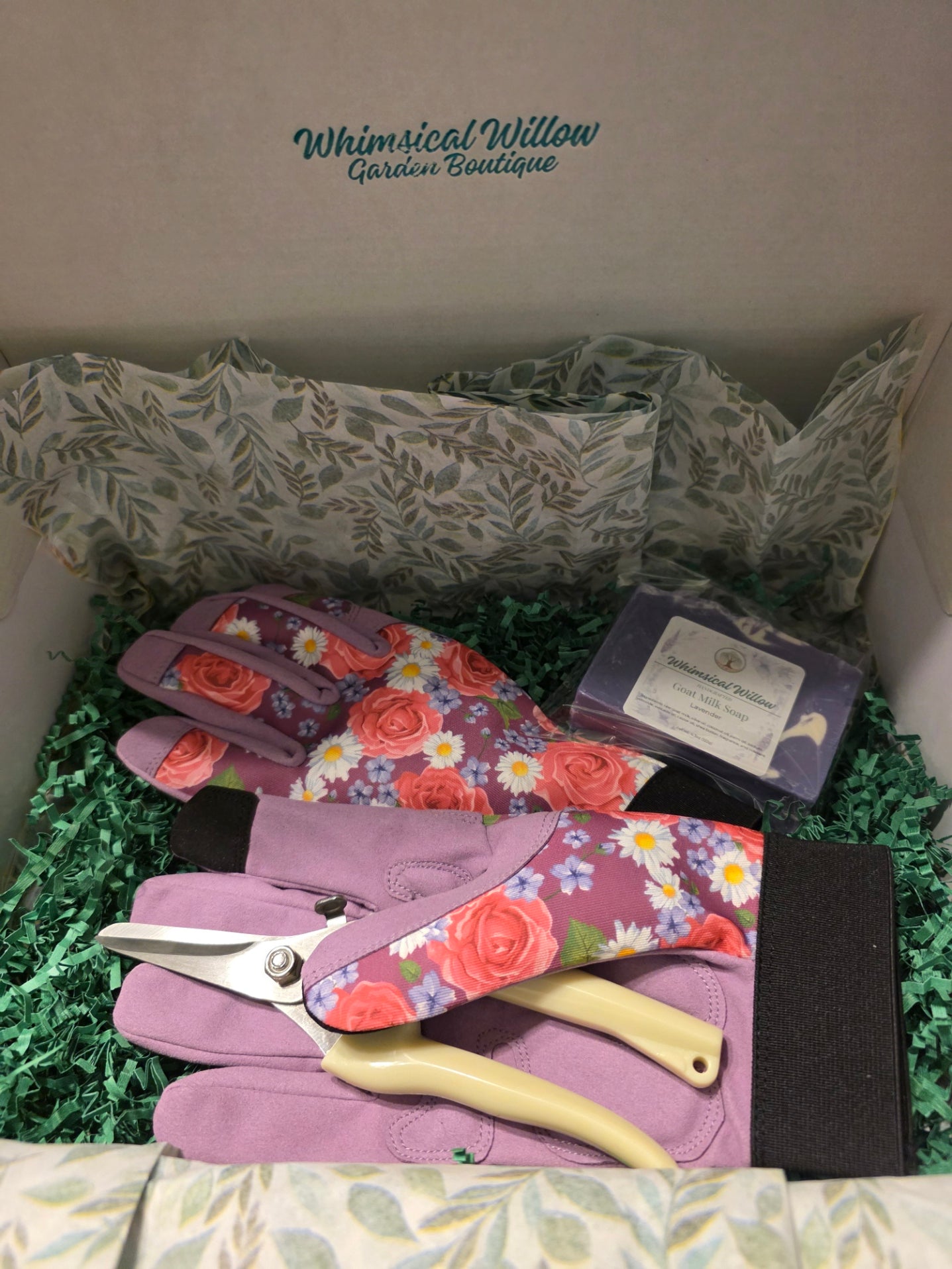 A gift box from Whimsical Willow Garden Boutique containing gardening gloves with a pink and purple rose floral print, gardening shears with cream handles, and a small packaged item, all arranged in green crinkle paper.