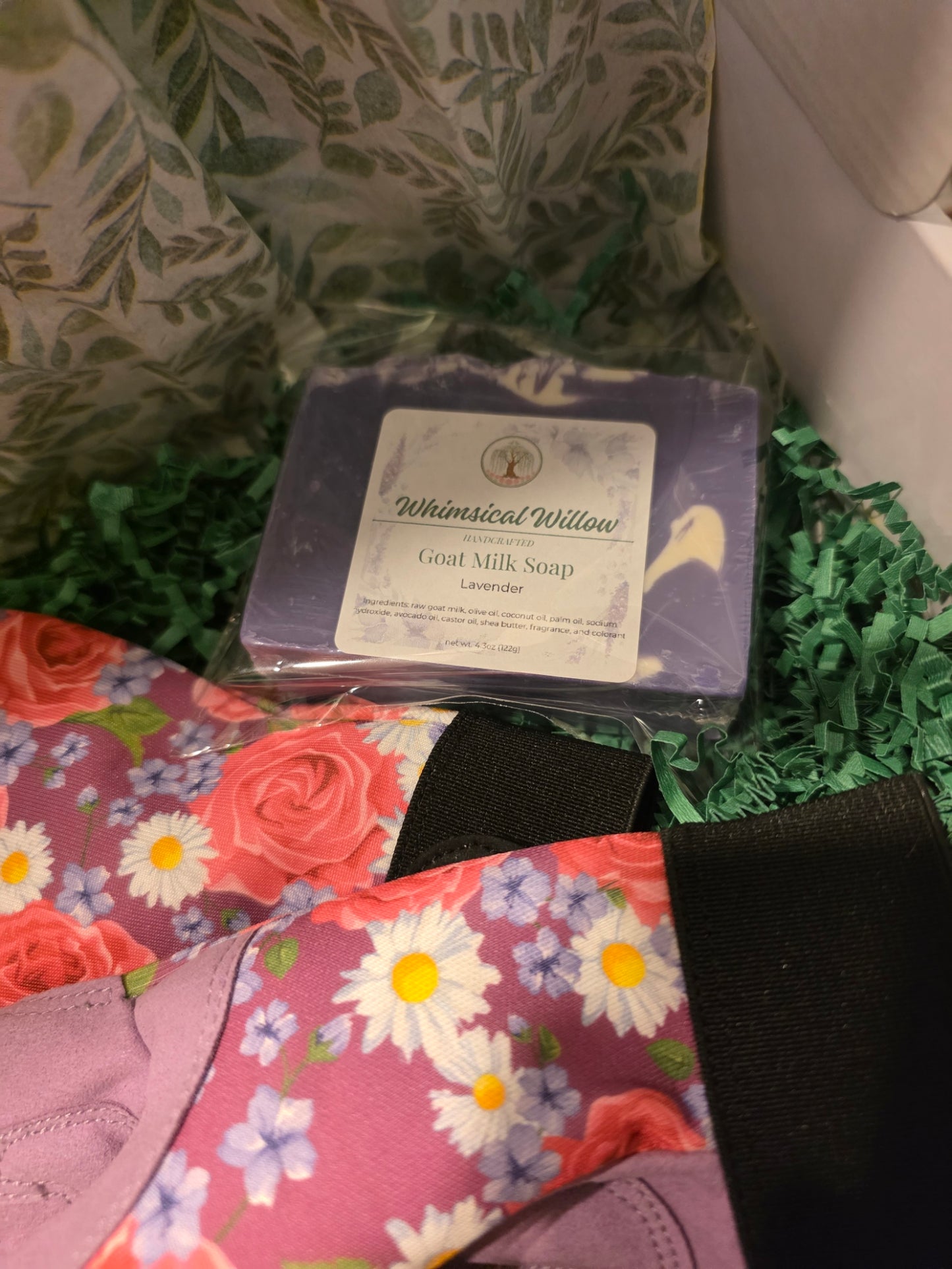 A packaged bar of soap labeled 'Whimsical Willow Great Milk Soap Lavender' resting on green paper filler inside a box, surrounded by floral fabric and a black strap.