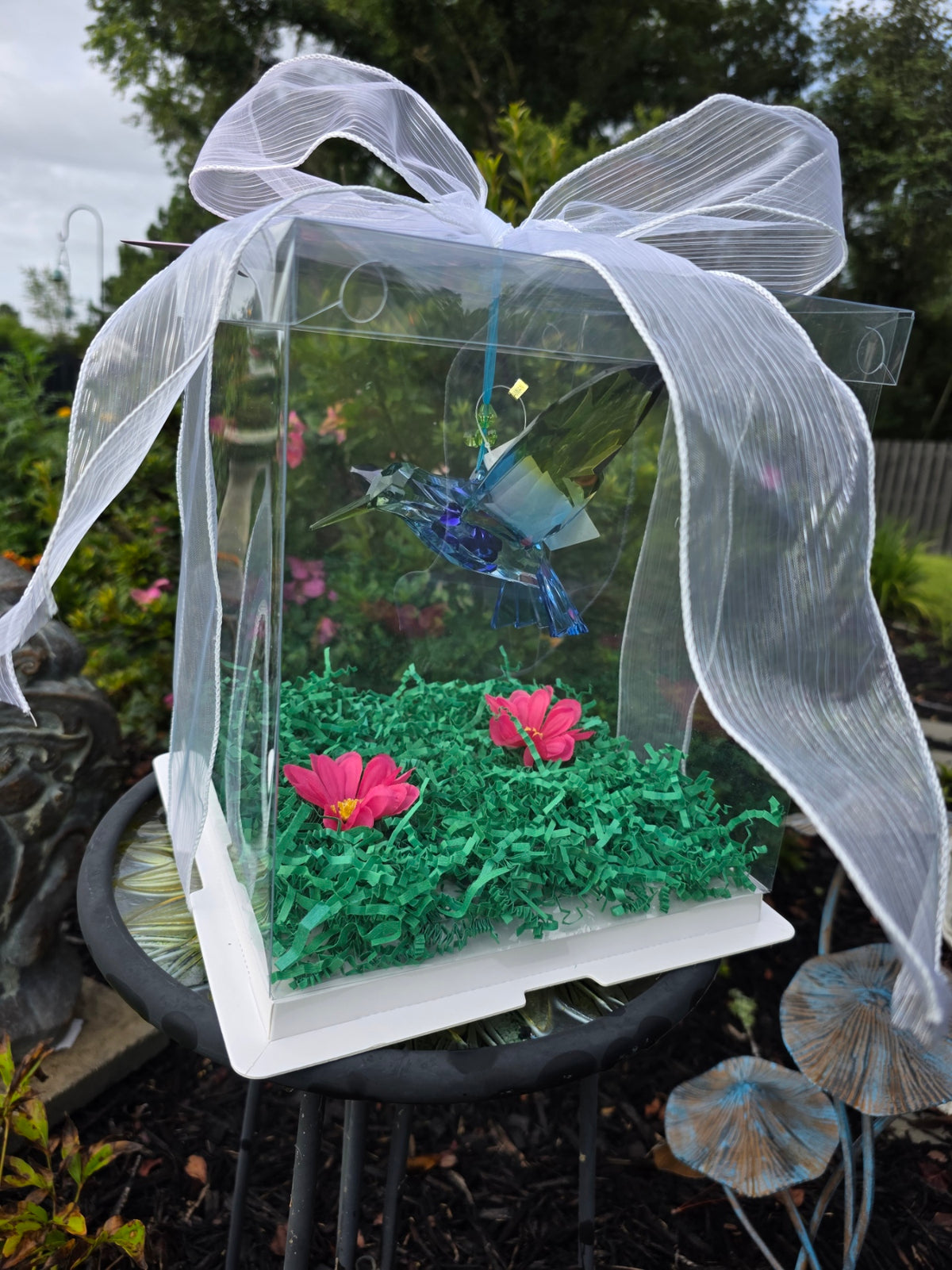 A clear acrylic gift box with a white base, tied with a large white organza bow, containing green shredded paper, two pink flowers, and a blue dragonfly decoration, photographed outdoors on a garden table.