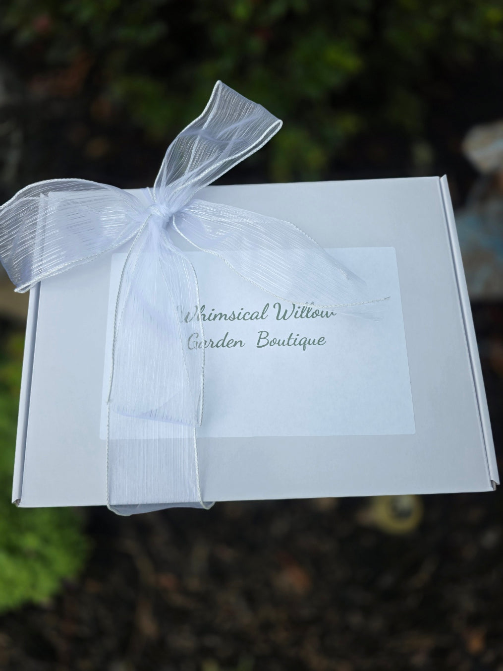 A white rectangular gift box tied with a sheer white ribbon bow, featuring a small white card with the text 'Whimsical Willow Garden Boutique' in gray script, set against a blurred dark green outdoor background.