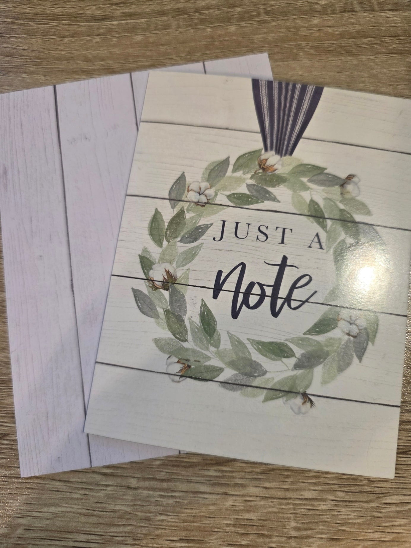 A square greeting card with a rustic white wood-plank design, featuring a green leaf wreath with white flowers and the words 'JUST A note' in black script. A matching white card or envelope is partially visible behind it on a wood surface.