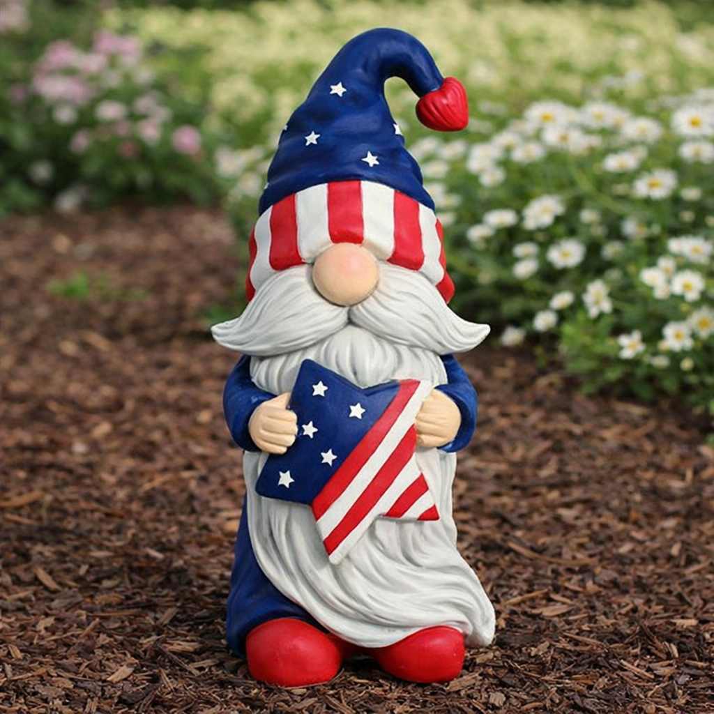 Gnome figurine with American flag hat and flag, standing on mulch with flowers in the background