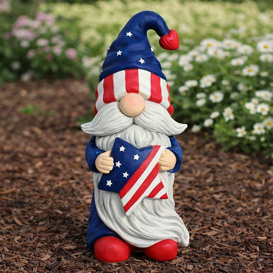 Gnome figurine with American flag hat and flag, standing on mulch with flowers in the background