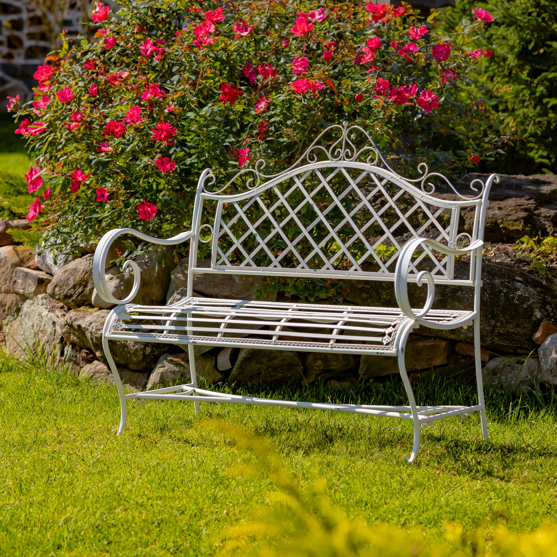 White metal garden bench in a garden setting with pink flowers and rocks.