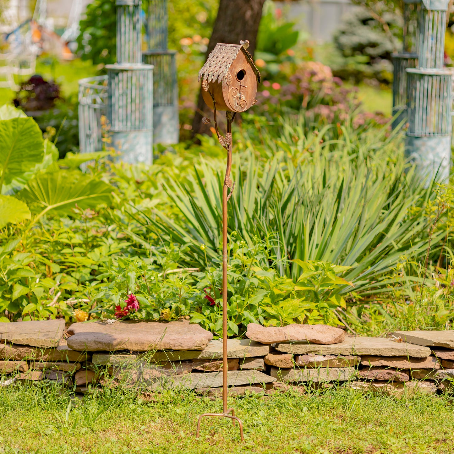 Decorative birdhouse on a stand in a garden setting with greenery and a stone wall.