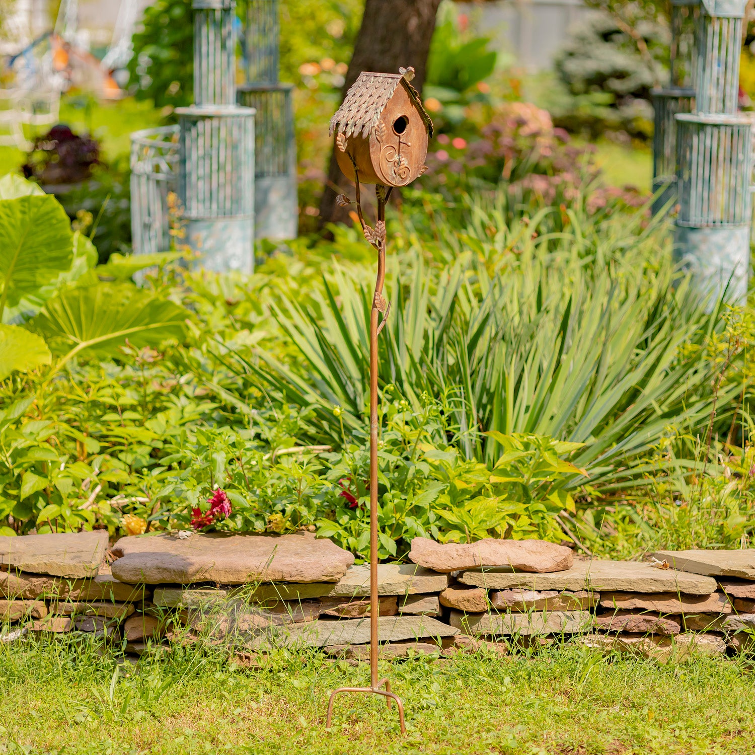 Decorative birdhouse on a stand in a garden setting with greenery and a stone wall.