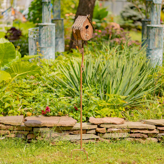 Decorative birdhouse on a stand in a garden setting with greenery and a stone wall.