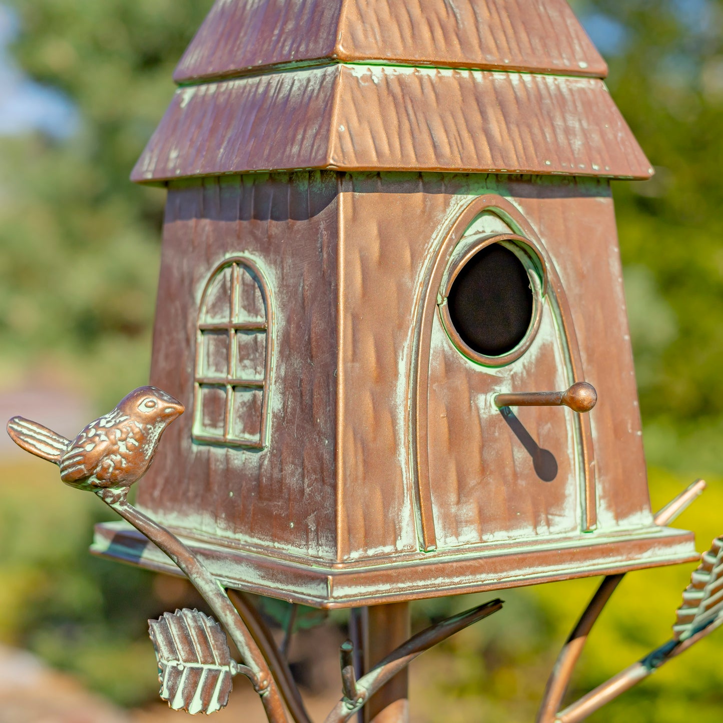 Bronze birdhouse with a bird perched on a branch against a blurred green background