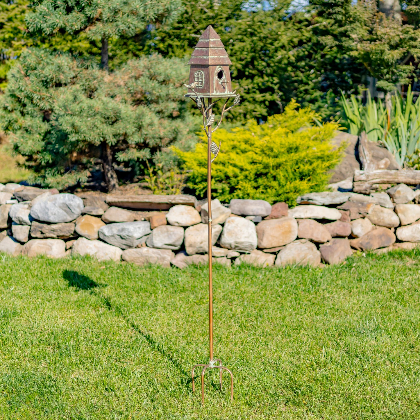 Decorative birdhouse on a stand in a garden setting with a stone wall and trees in the background.