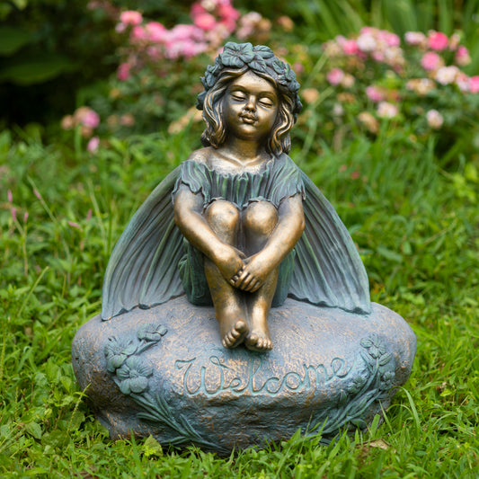 Bronze fairy statue on a stone base with 'Welcome' engraved, set against a garden backdrop.