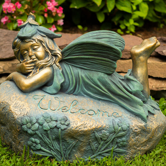 Decorative fairy statue on a rock with 'Welcome' inscription, surrounded by greenery.