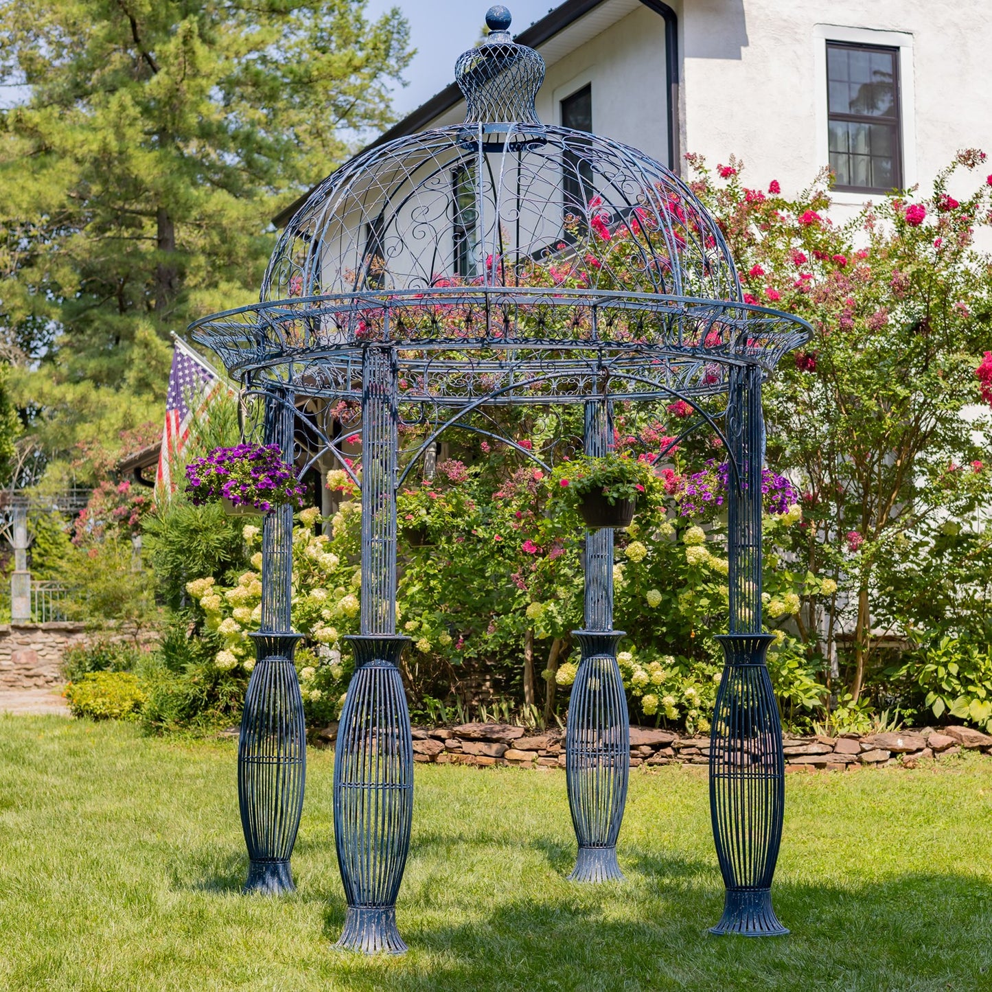 A large decorative metal garden gazebo with a domed top and four fluted legs, shown outdoors on a lawn in front of flowering bushes and a house.