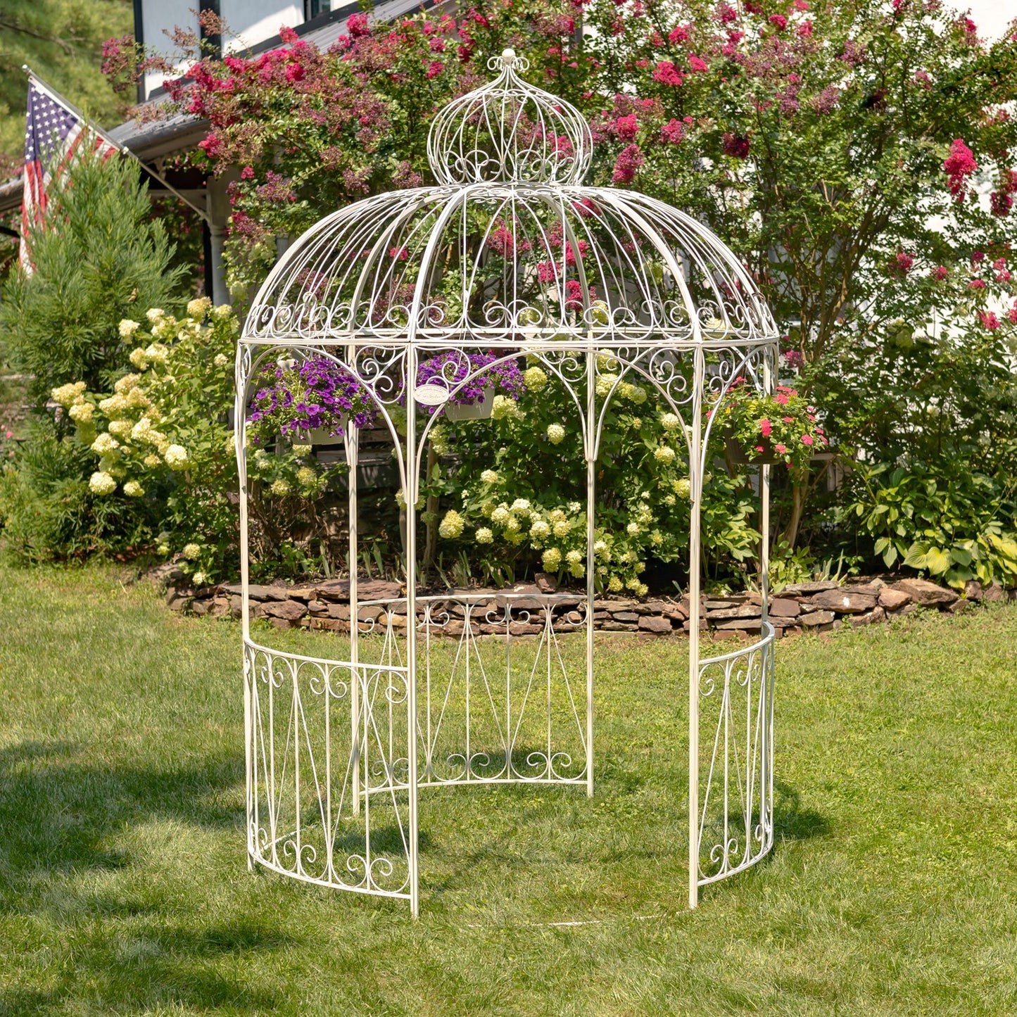 A white wrought-iron garden gazebo with a domed roof and decorative scrollwork, set on a green lawn in a landscaped garden with pink and purple flowers.