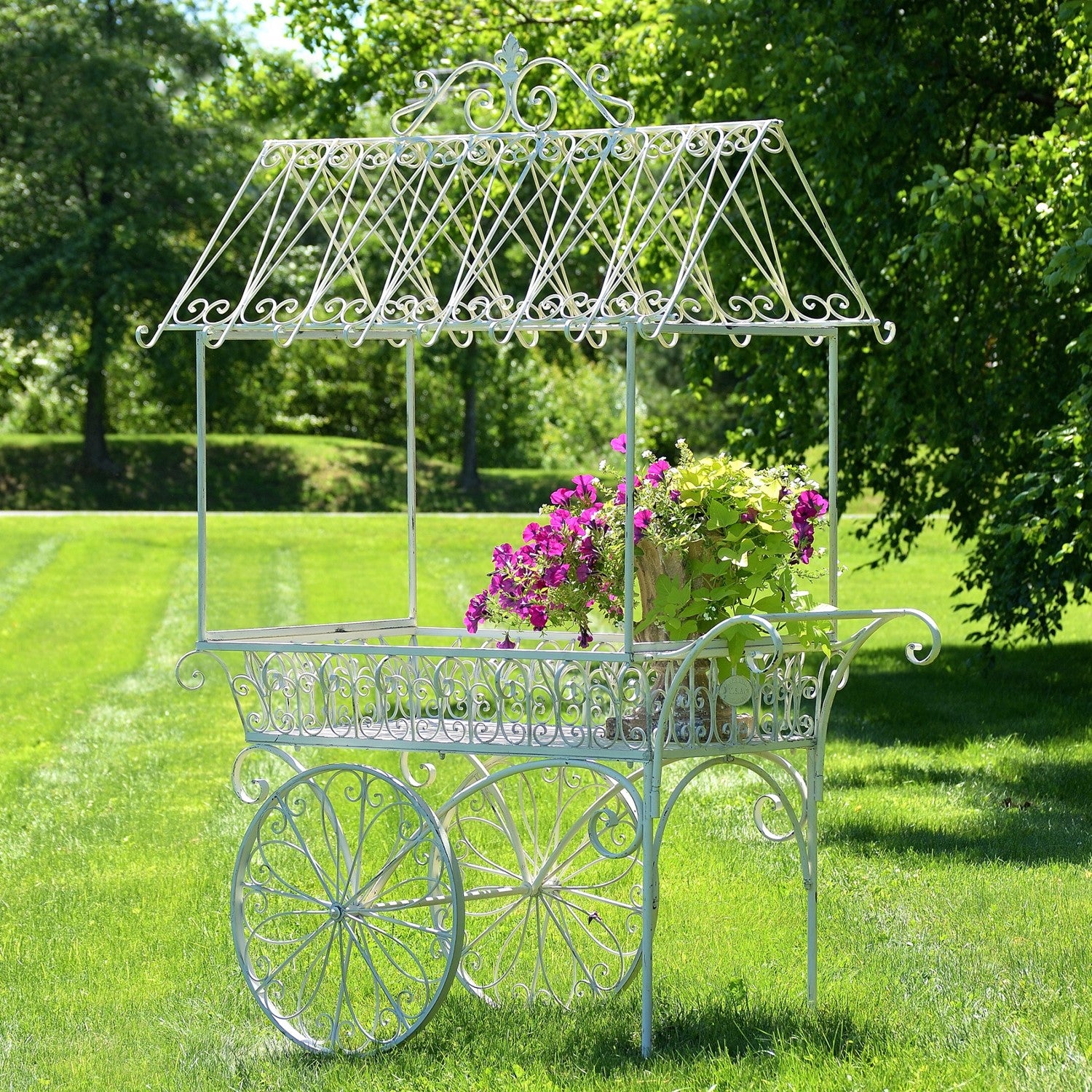 A decorative white wrought-iron garden cart with ornate scrollwork and four wheels, placed on a green lawn outdoors with trees in the background. A potted plant with purple flowers sits on the cart.