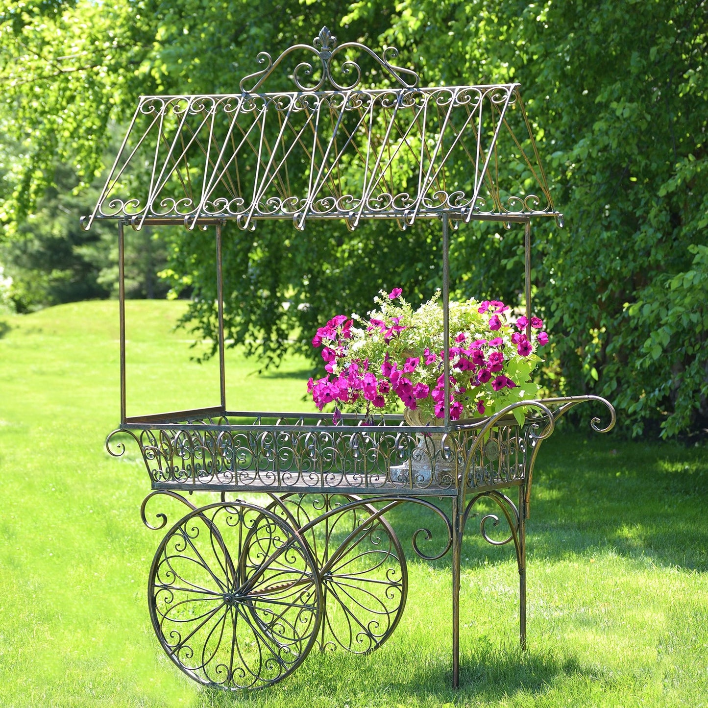 An ornate vintage-style metal flower cart with a canopy and large spoked wheels, placed on a grassy lawn with a lush green garden background. A potted plant with bright pink and white flowers is placed inside the cart.