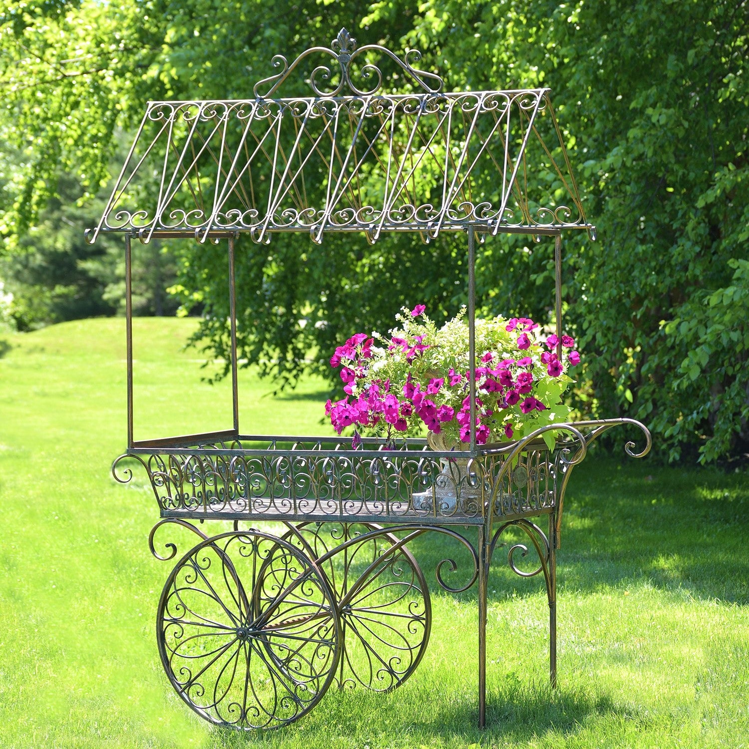 An ornate vintage-style metal flower cart with a canopy and large spoked wheels, placed on a grassy lawn with a lush green garden background. A potted plant with bright pink and white flowers is placed inside the cart.