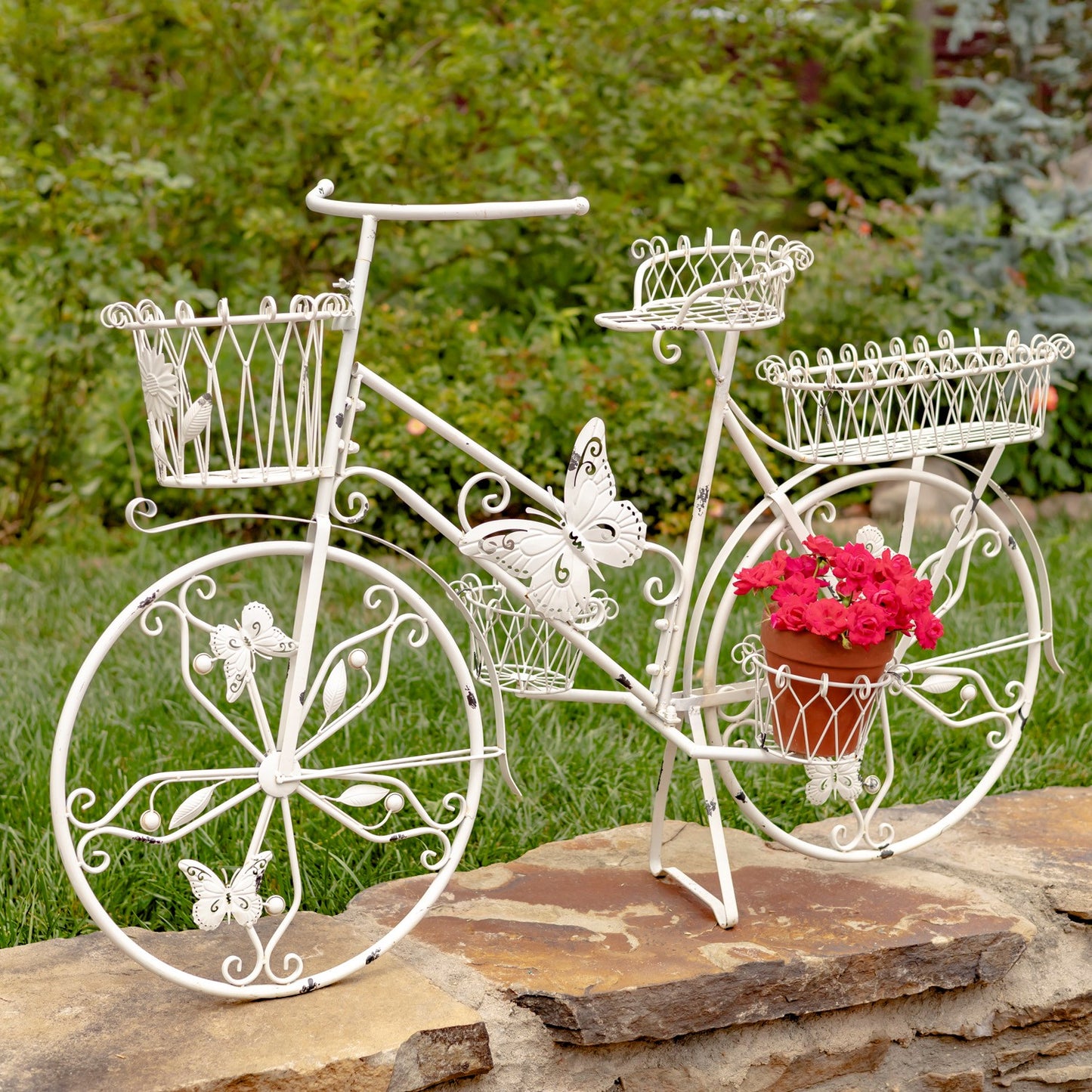 A white metal decorative garden bicycle sculpture with butterfly details and wire baskets, displayed outdoors on a stone surface with green grass and bushes in the background.