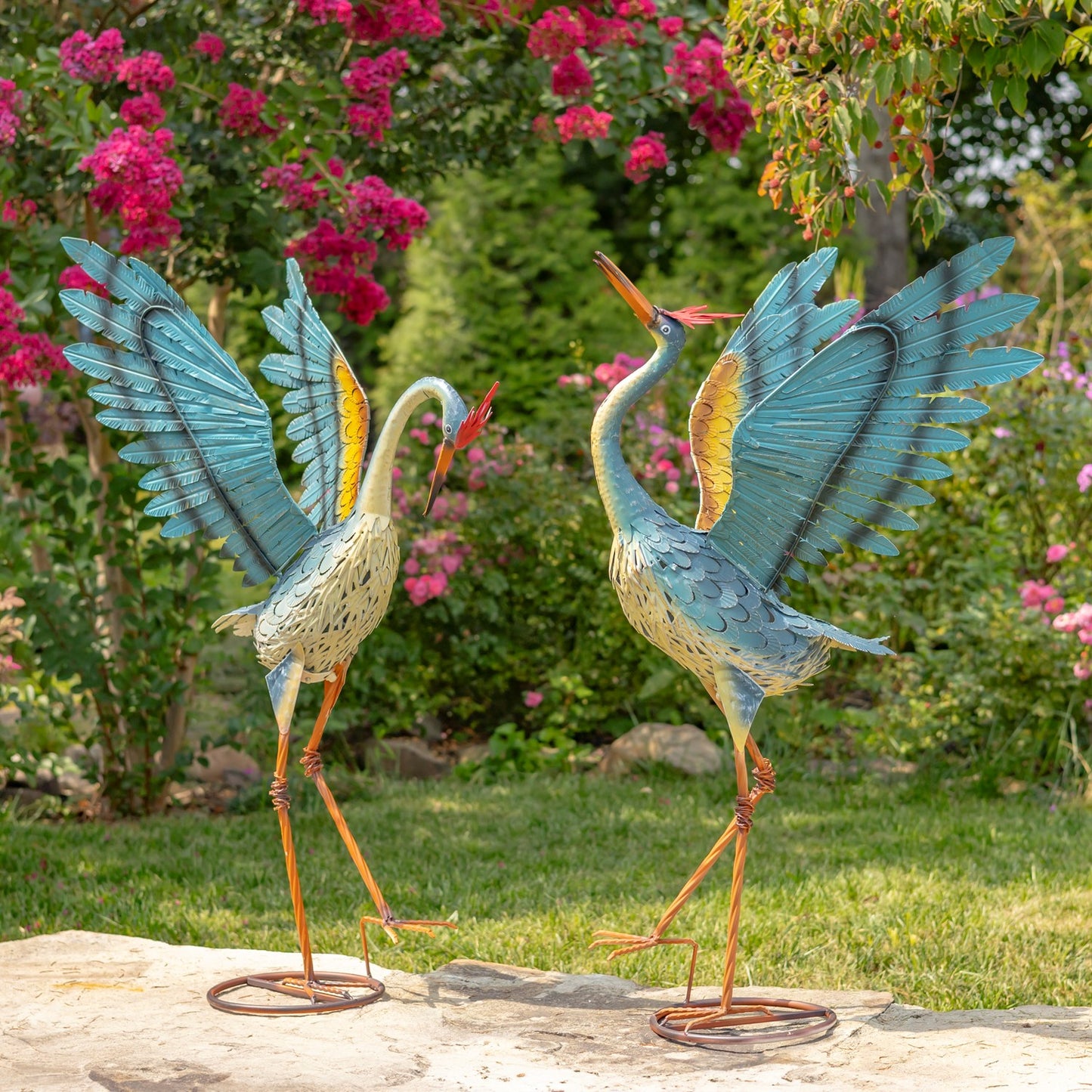 Two decorative garden statues of herons with spread blue wings and orange legs, displayed on a stone surface in a garden with pink flowers and green grass.