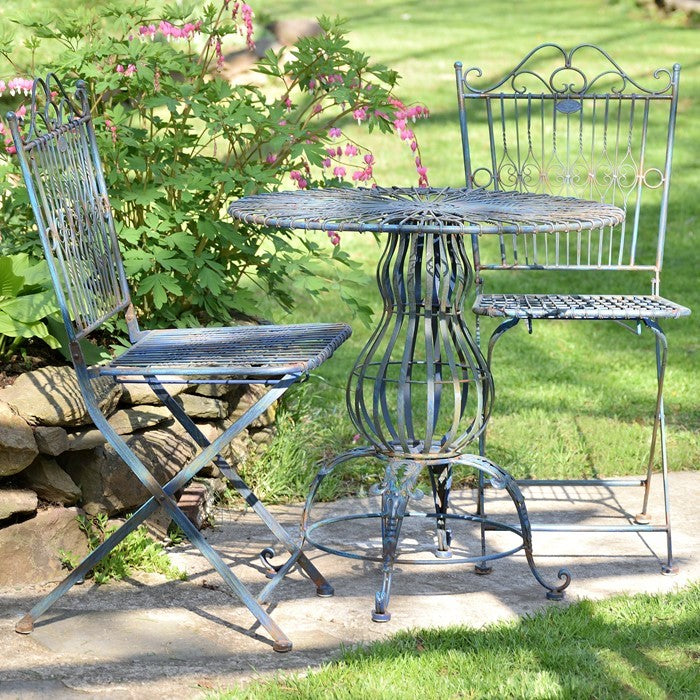 A three-piece metal bistro set with a round table and two chairs in a garden setting. The set is painted in a distressed blue-gray finish, featuring a wire mesh table top and seat, and an openwork metal chair back and legs.