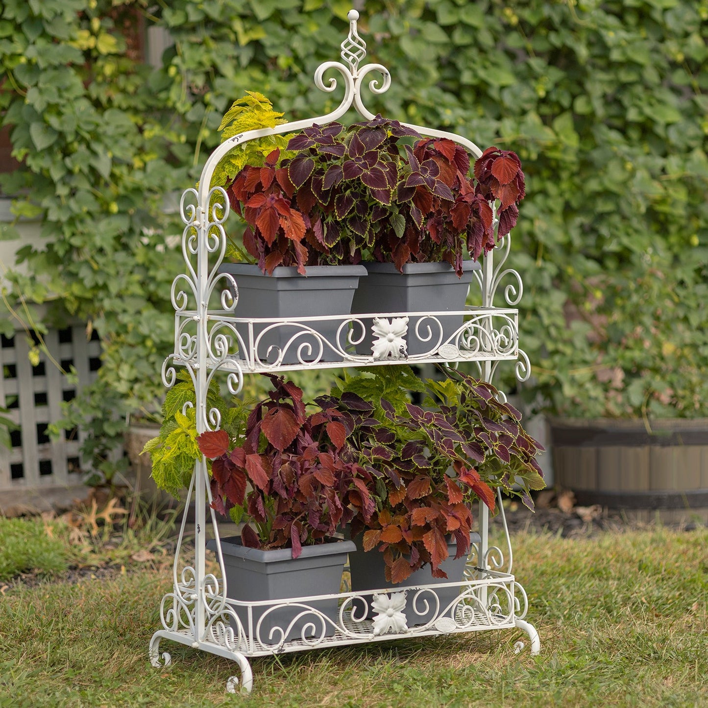 Antique white two-tier metal plant stand with decorative scrollwork displaying potted flowers on a cottage-style porch or garden patio.