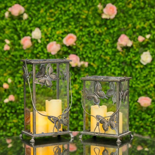 Two decorative metal lanterns with butterfly and leaf motifs, each holding a yellow pillar candle, displayed on a reflective surface against a blurred green foliage background with pink flowers.
