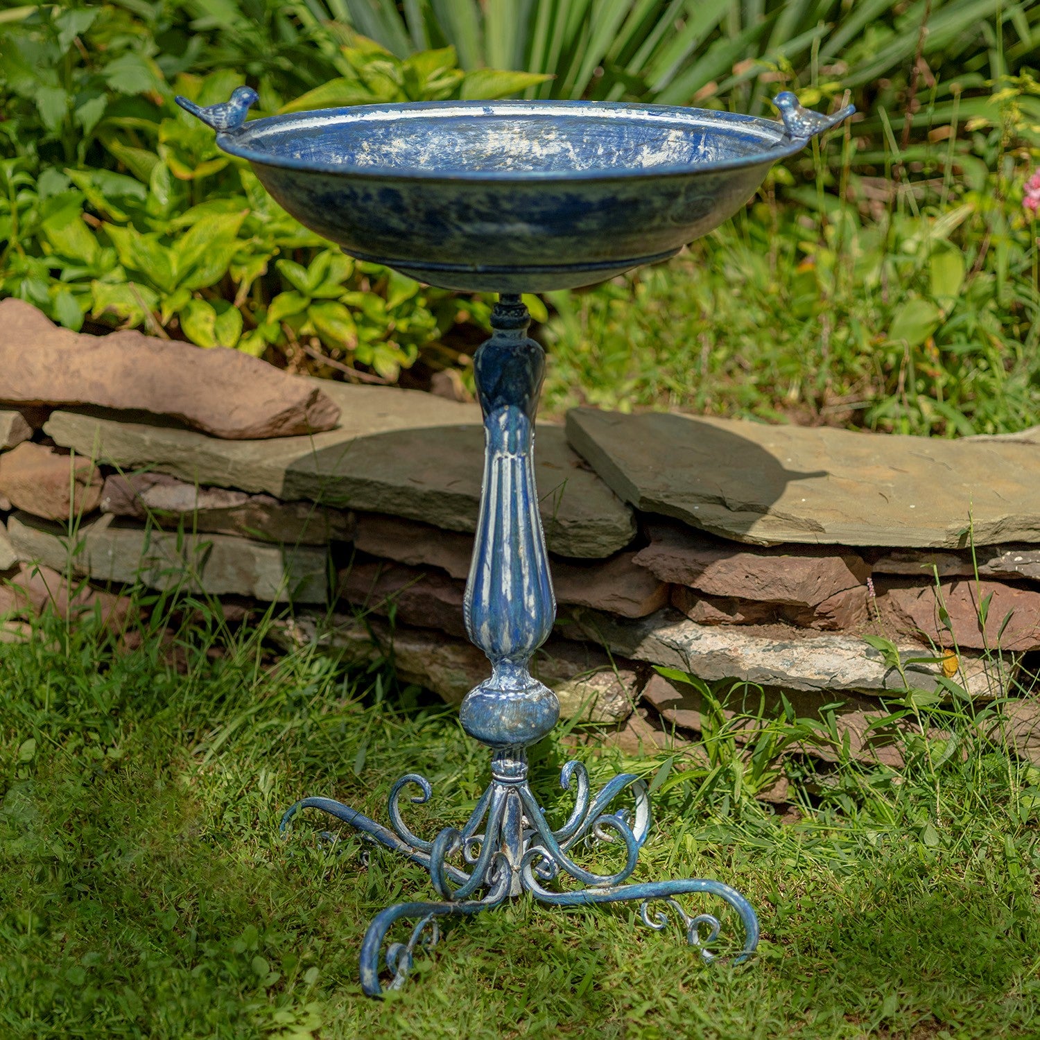 Decorative blue metal bird bath in a garden setting with greenery and a stone wall.