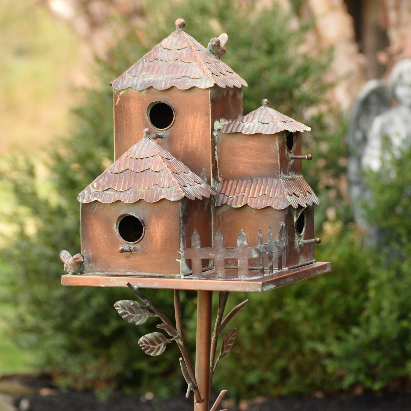 A decorative copper birdhouse with three tiered roofs and bird perches, mounted on a metal stake with leaf details, set against a blurred garden background.
