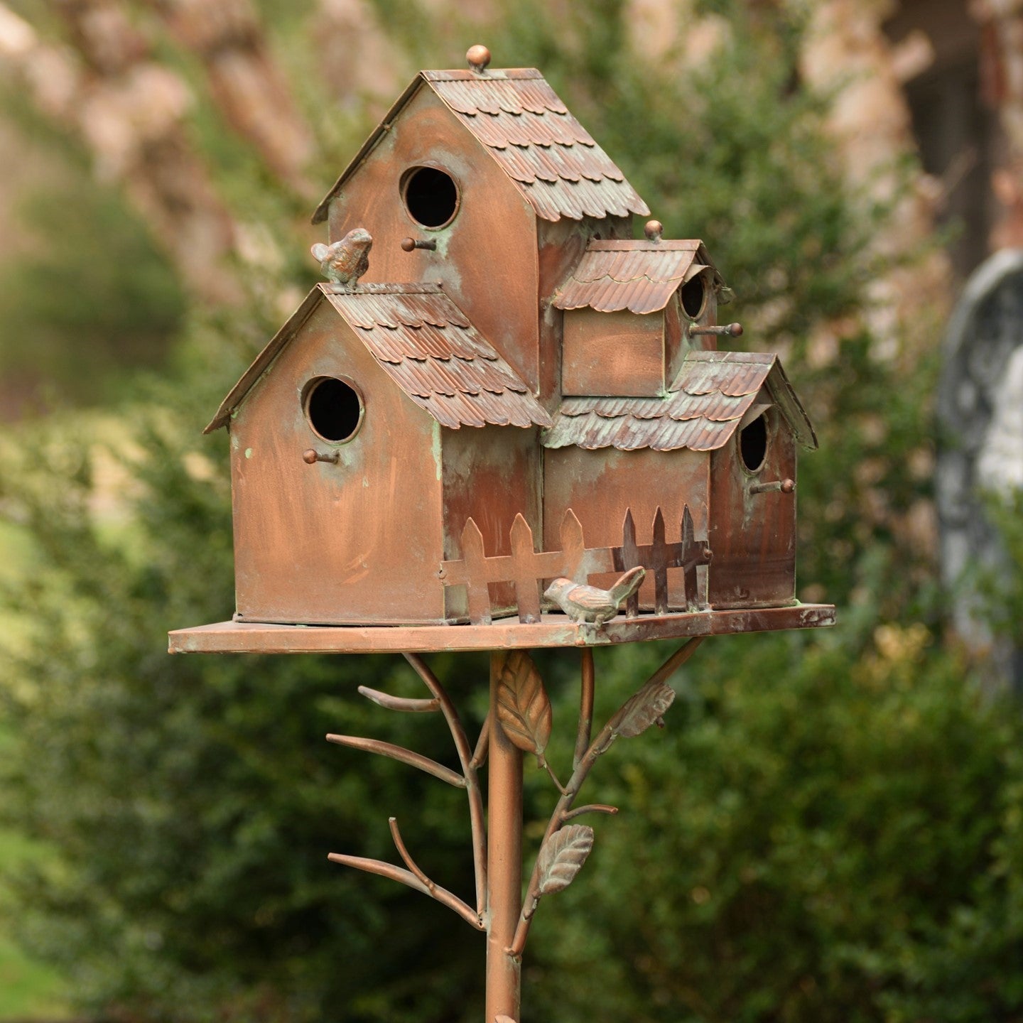 A rustic multi-roof birdhouse with a weathered copper-brown finish, mounted on a decorative metal stake shaped like branches, set against a blurred garden background.