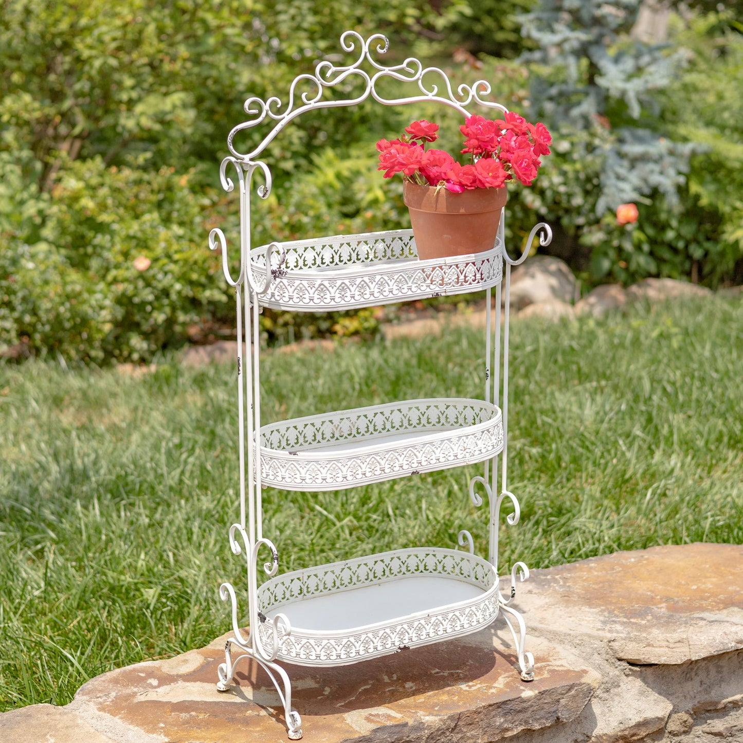 A three-tier white metal garden stand with decorative scrollwork and cutout patterns, holding a potted red flower, set outdoors on a stone surface with a green lawn and garden in the background.