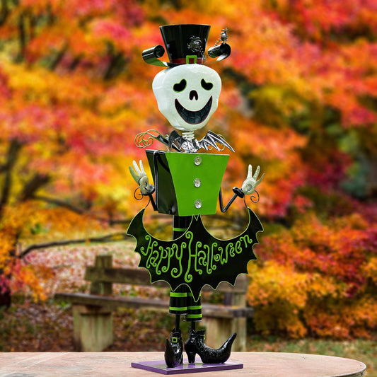 A decorative metal Halloween figurine of a skeleton character wearing a black top hat and bow tie, holding a green box with silver buttons, and a black bat-wing shaped sign reading 'Happy Halloween' in green. The figurine stands on a purple base against a blurred autumn park background with orange and red foliage.