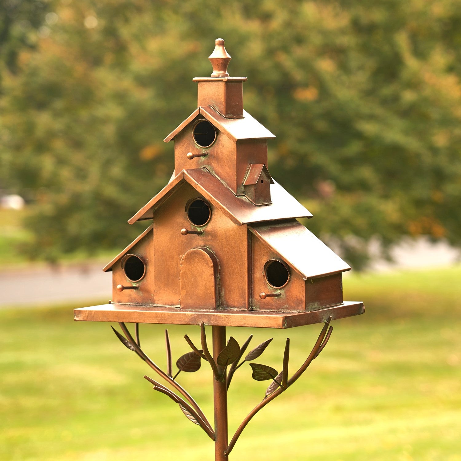 A rustic metal birdhouse with a tiered cottage-style roof and three circular entry holes, mounted on a decorative branch-shaped stake in an outdoor garden setting.