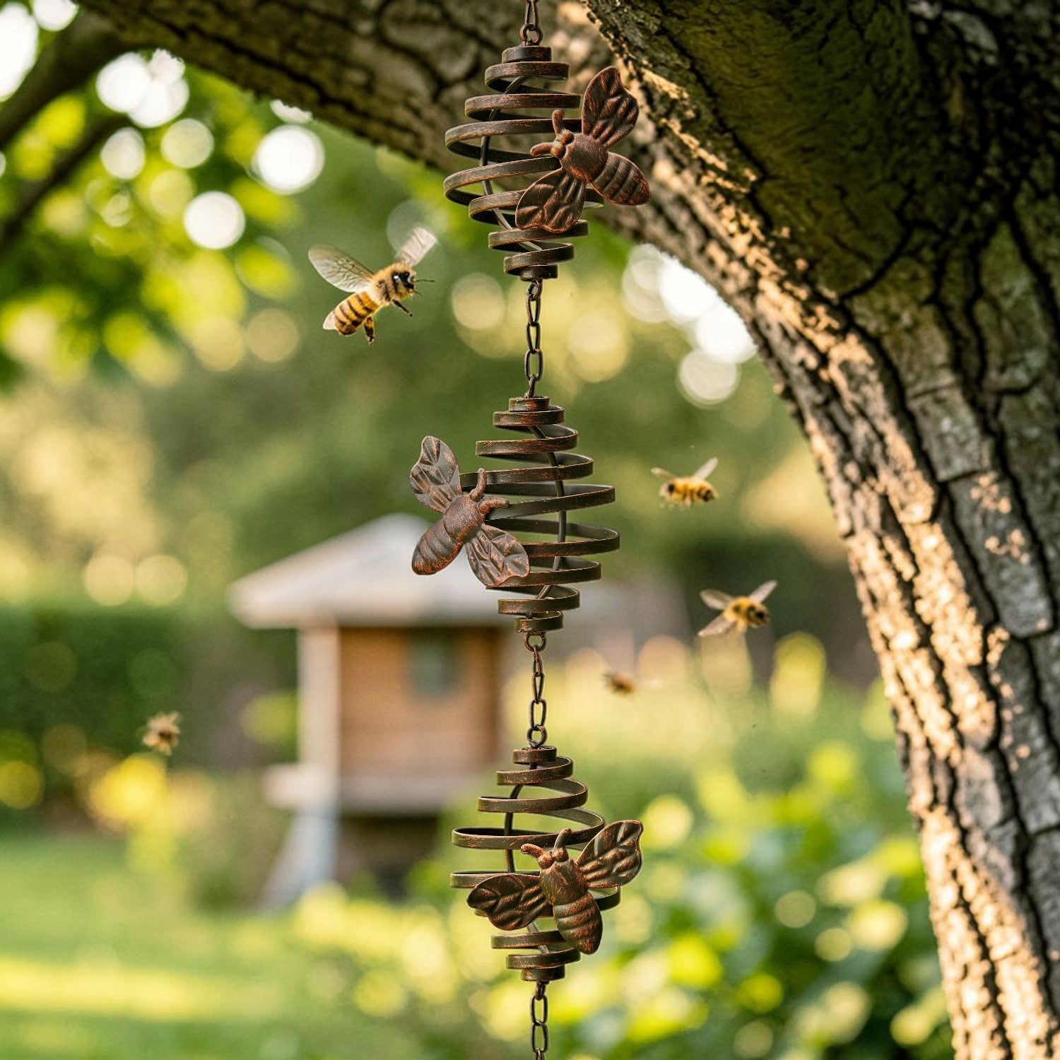A rustic metal bee wind spinner hanging from a tree branch in a garden, with several flying bees and a blurred birdhouse in the background.