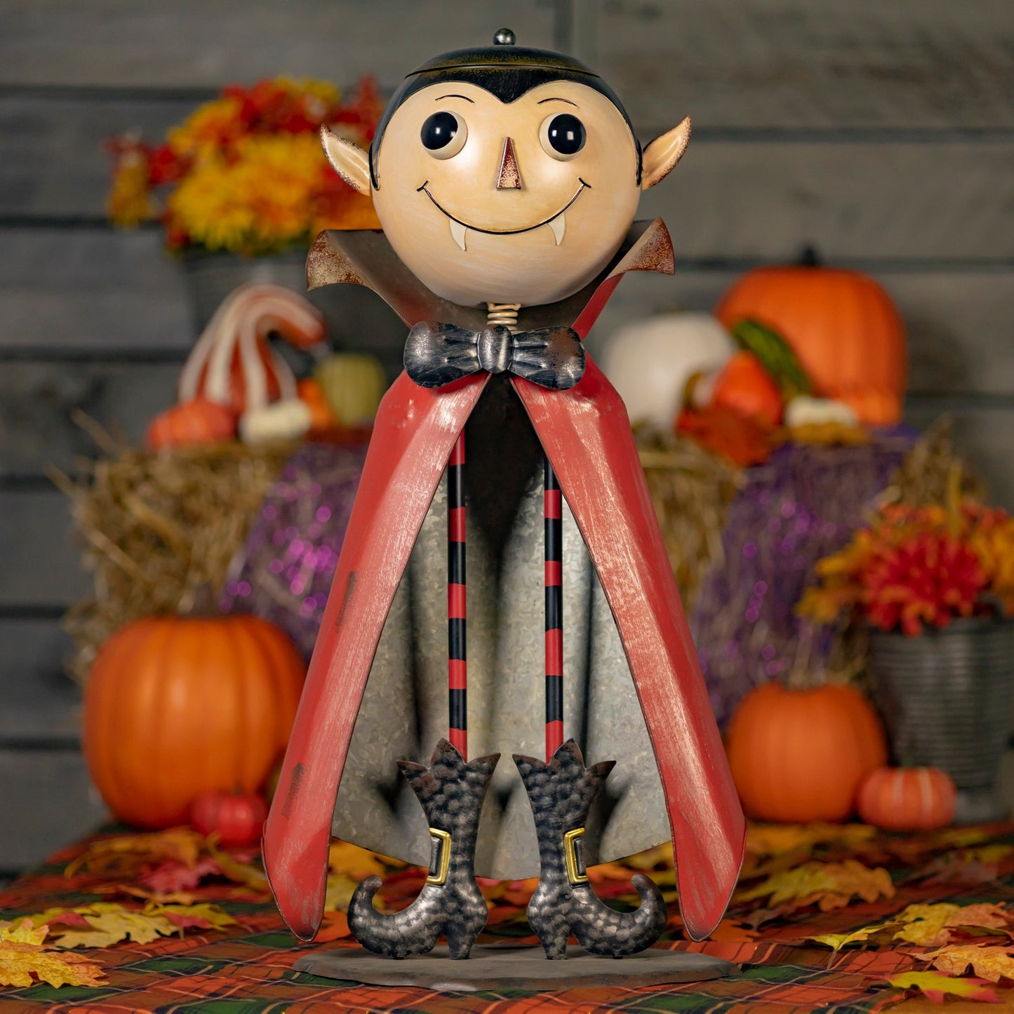 A decorative vampire figurine with a round beige face, pointed ears, black eyes, and fangs, wearing a red cape, black-and-red striped legs, and clawed shoes, posed among pumpkins, autumn leaves, and fall flowers.
