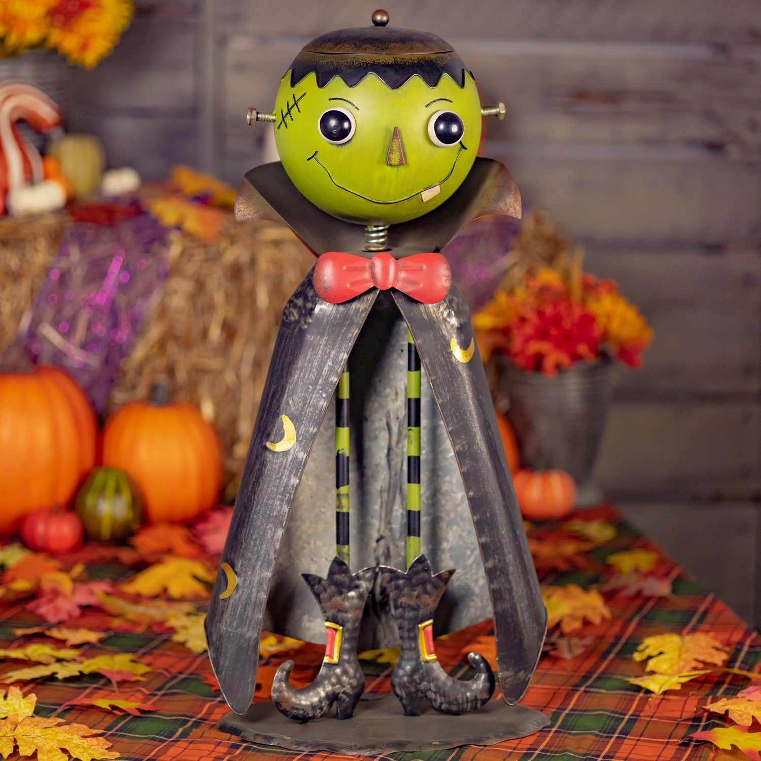 A Halloween decorative metal figure shaped like Frankenstein's monster, standing on a plaid autumn table runner with pumpkins and fall leaves in the background. The figure has a green head, black hair, a pink bow tie, a dark gray cape with yellow crescent moon details, and black witch boots.