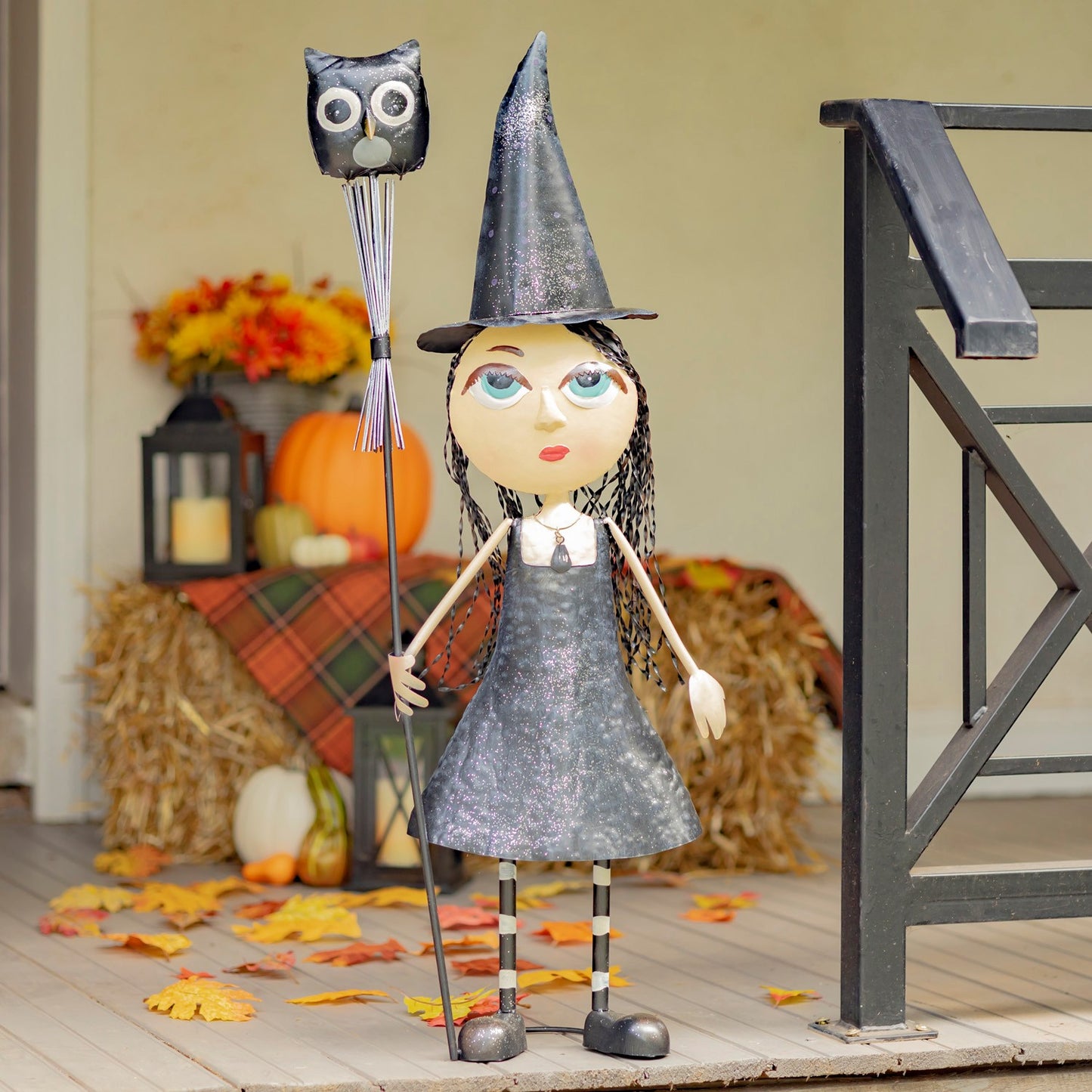 A decorative witch doll figurine with a glittery gray dress, black pointed hat, and black-and-white striped legs, holding a wand topped with an owl head. The doll is posed on a porch-style backdrop with pumpkins, hay, autumn leaves, and a railing.