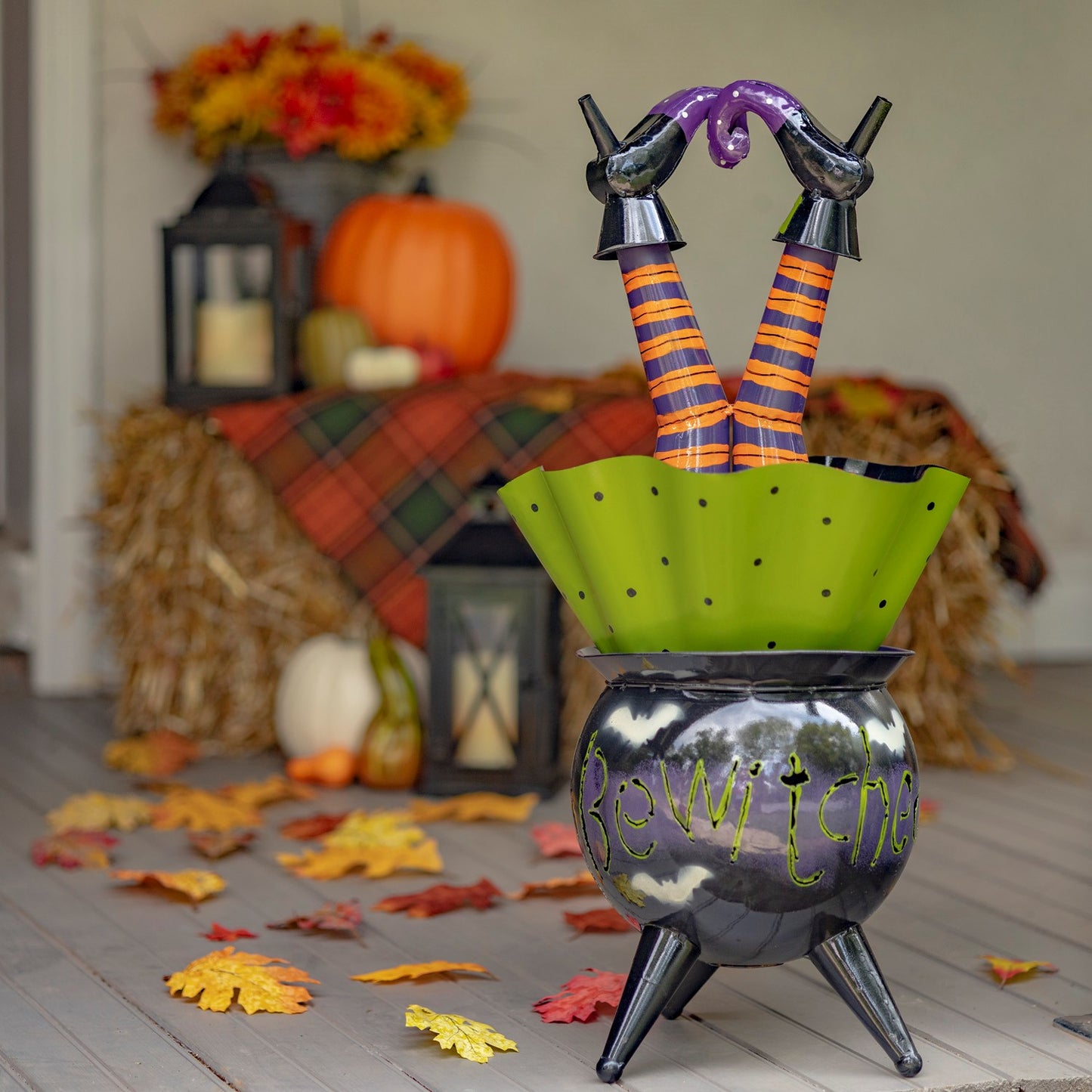 A Halloween decorative witch legs standee in a cauldron, featuring striped orange and purple legs, a green polka-dot skirt, and black shoes, set on a gray wooden floor with autumn leaves and fall decorations in the background.