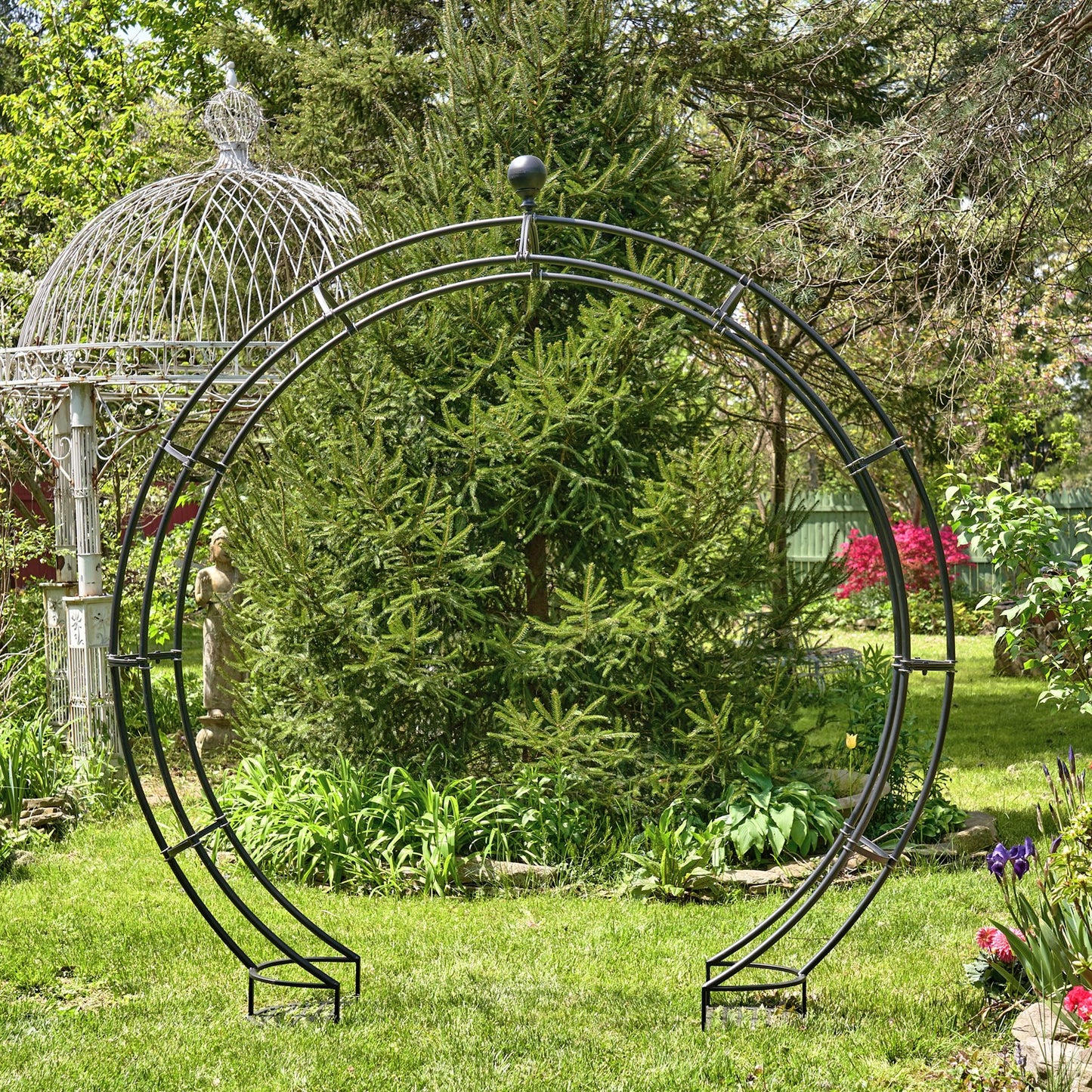 Circular metal garden archway in a lush garden setting with trees and flowers.